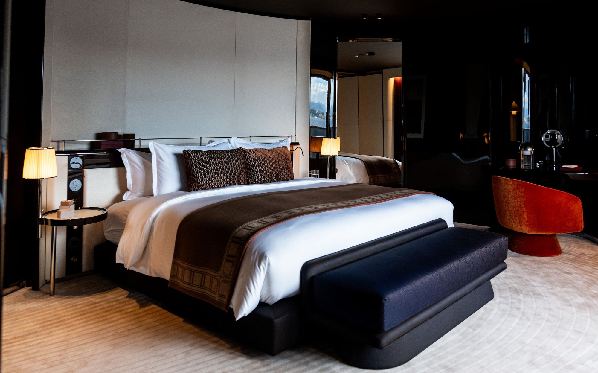 A luxury modern bedroom in a yacht featuring a large bed dressed in white linens with dark brown patterned accents cushions, set against with bedside tables and lamps. A dark upholstered bench sits at the foot of the bed, and a vibrant orange-red accent chair is visible in the background.