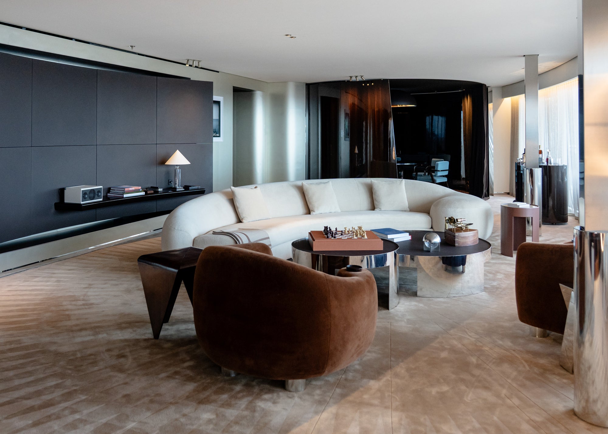 A modern living room interior featuring a curved cream sectional sofa, two plush brown armchairs, round dark coffee tables, and a large beige patterned area rug. The room has a dark gray accent wall on the left, recessed ceiling lights, and a glass-paneled doorway in the background.
