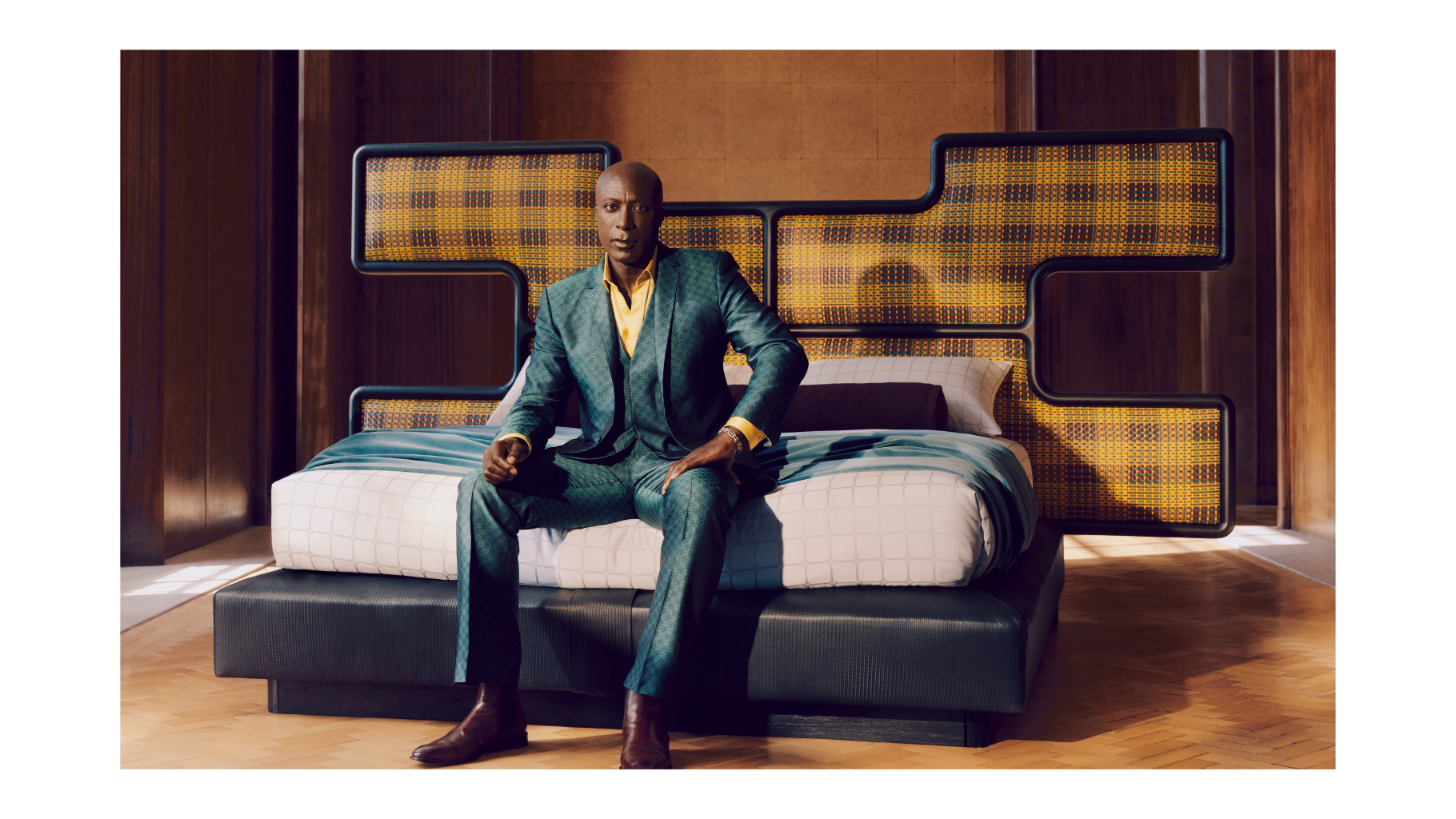 Ozwald Boateng in a suit sitting on a modern bed with a kente fabric headboard.