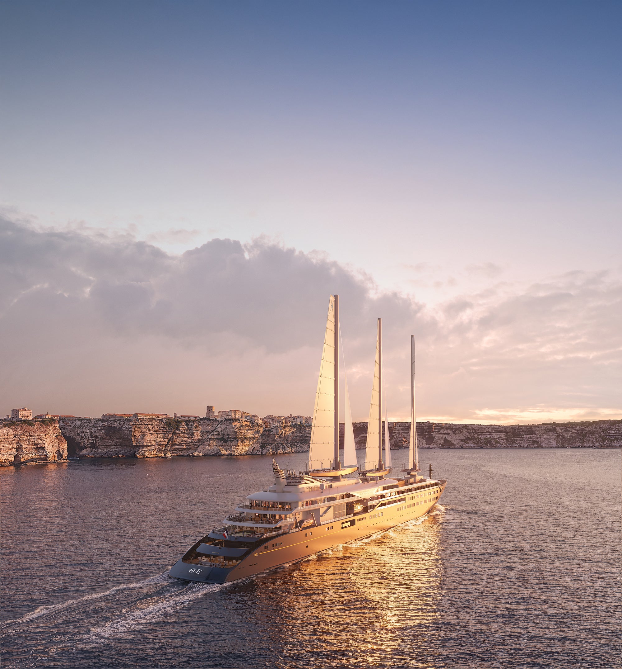 The Corinthian a large luxury yacht with tall white sails glides across calm blue water toward a rocky coastline at sunset, with clouds in the sky and warm light reflecting on the water.