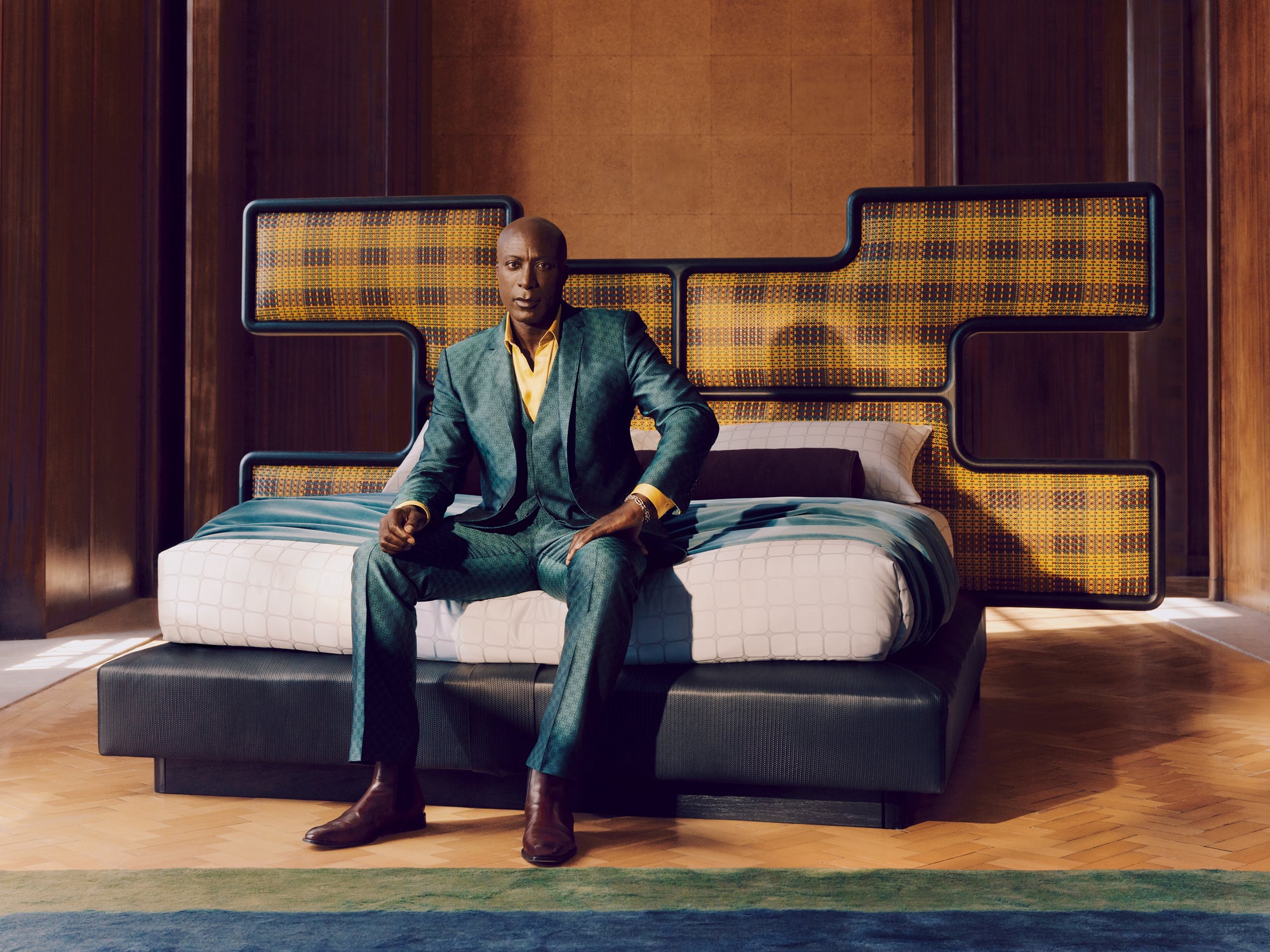 The Saville row Ozwald Boateng in a suit sitting on a modern bed with geometric headboard