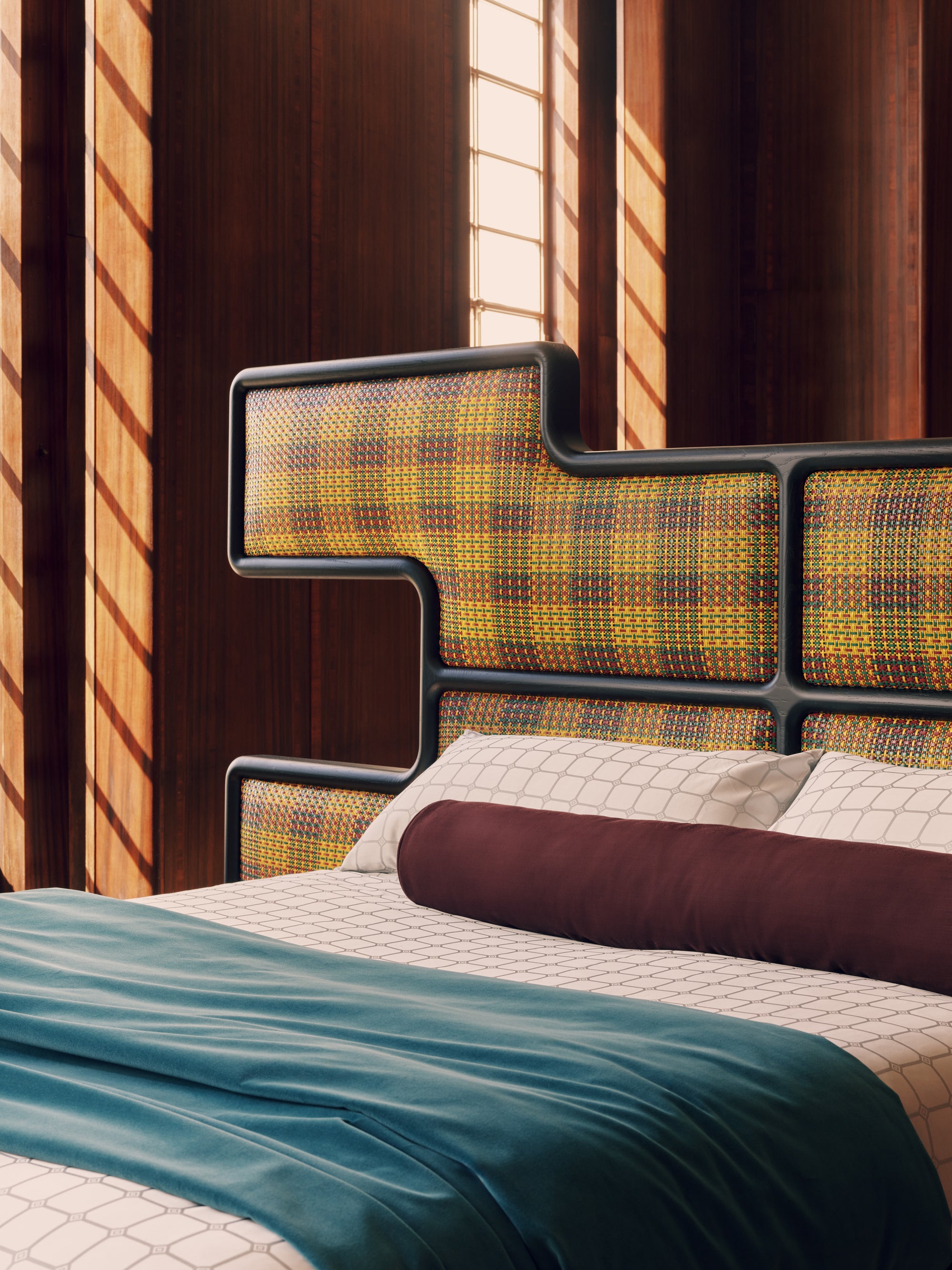 Modern bed with a kente fabric headboard in a room with wooden walls and a window.
