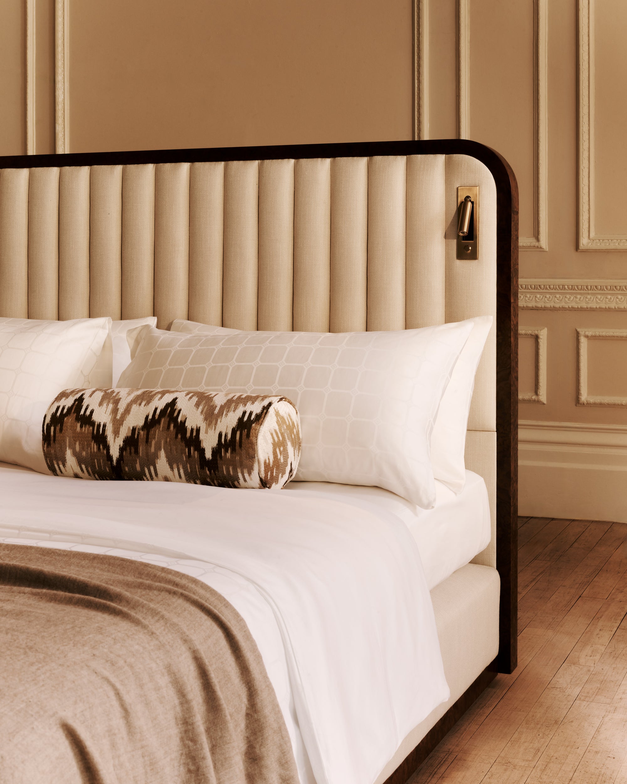 The Luna bed with a beige upholstered headboard with featuring white bedding and patterned pillow against a beige wall.