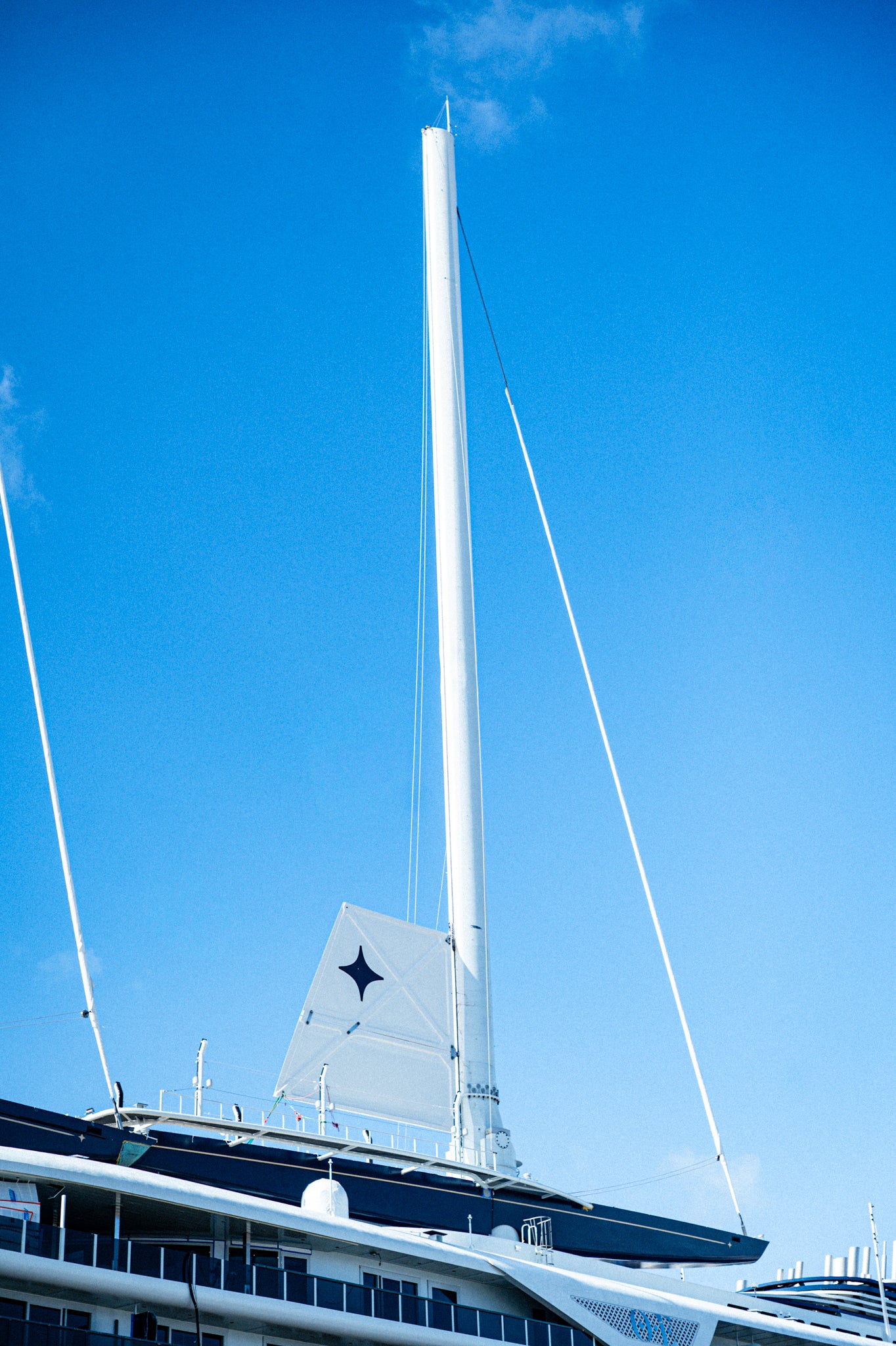 A close-up low-angle view of the orient express sailing yacht mast and rigging against a bright blue sky, with part of the dark blue and white vessel deck visible at the bottom.